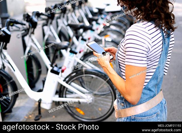 Woman standing near bicycle parking stand using smart phone in city