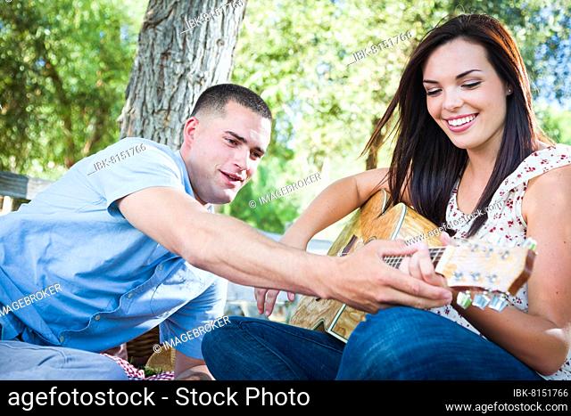 Young adult man teach his girlfriend how to play the guitar outside in the park