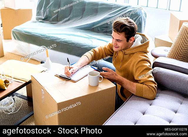 Smiling man with mobile phone writing on paper over cardboard box in new home