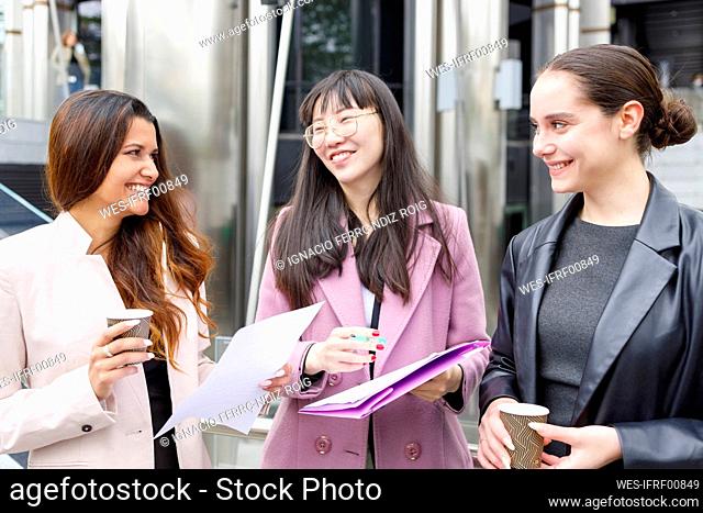 Smiling businesswomen discussing while standing with coffee cups and files