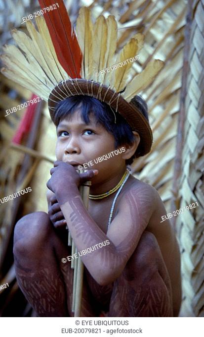 Young boy playing panpipes outside maloca prior to a festival. Painted with deep purple we leaf dye and wearing crown of red macaw and yellow toucan feathers