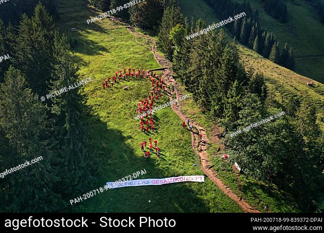 18 July 2020, Bavaria, Rettenberg: Around 80 people dressed in red stand on a mountain meadow on the Grünten in the form of a question mark above a poster with...