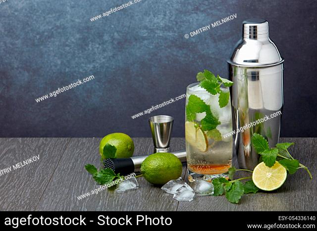 Set of bar tools: cocktail shaker, muddler, jigger, cocktail with ice in highball glass, lime and mint on dark gray background with copy space