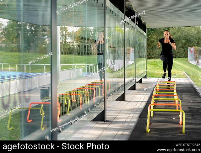 Sportswoman running over small hurdle by glass wall at ground