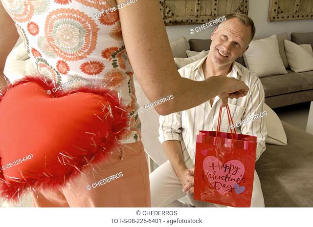 Mid section view of a woman hiding a stuffed red heart behind her back with a mid adult man sitting on a couch