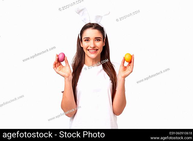 Portrait of lovely caucasian female in rabbit ears, showing two colored eggs and smiling, congratulate everyone with happy Easter day