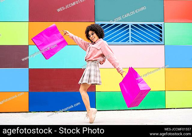 Cheerful woman with shopping bags standing on tiptoe by colorful wall