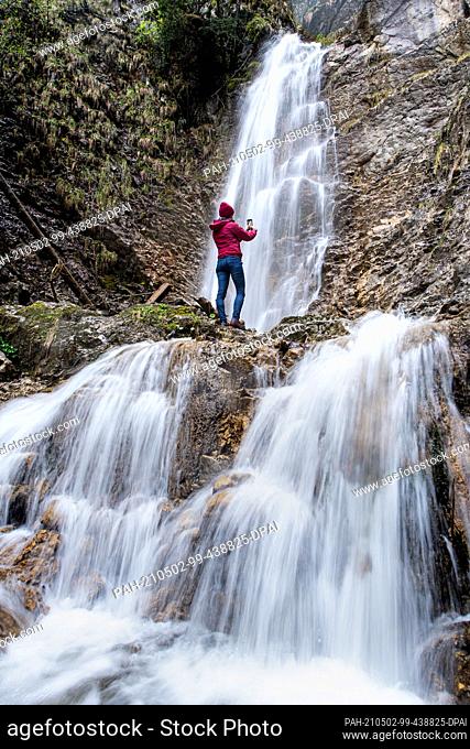 02 May 2021, Bavaria, Grassau: A woman stands at the Grießenbach waterfall in rainy weather and takes a photo with her smartphone