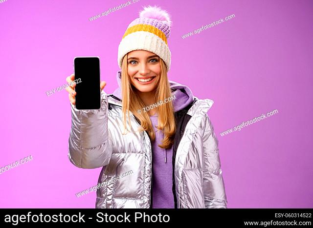 Lifestyle. Friendly cheerful confident blond girl in silver stylish winter jacket hat extend arm showing smartphone display advertising awesome new device app...