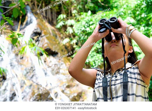 Beautiful young girl hiking is using binoculars look for birds in tropical forest near the waterfalls in Thailand