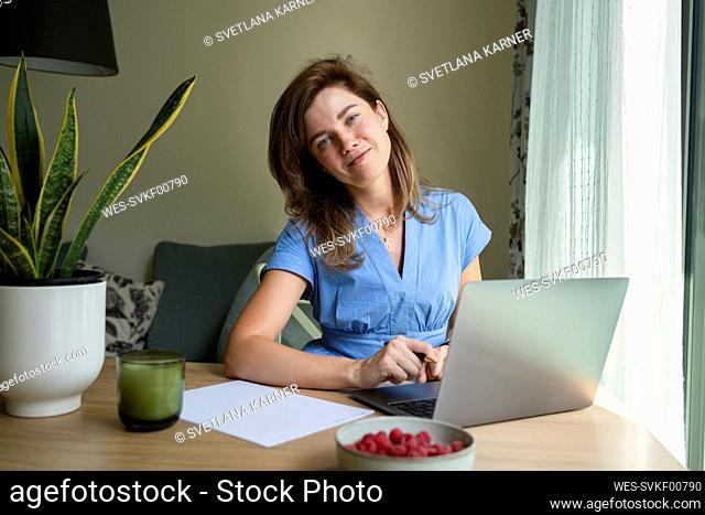 Smiling freelancer sitting with laptop at desk in home office