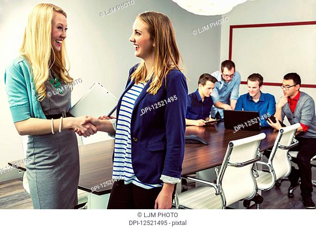 A young millennial business professional commending her employee on her efforts while her co-workers carry on their work in a conference room at their place of...