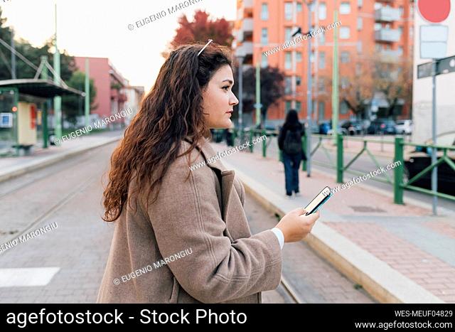 Young woman in overcoat looking away while holding mobile phone