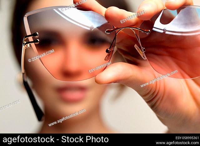 Portrait of young woman with sunglasses in hand. Female is looking at the camera through glasses. Focus on sunglasses and fingers, face is in bokeh
