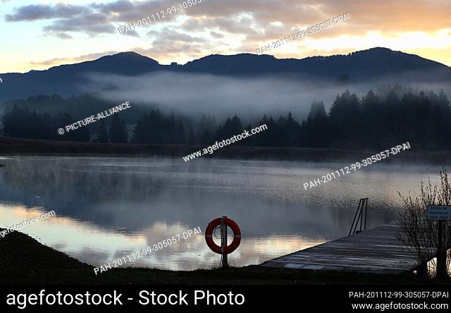 12 November 2020, Bavaria, Roßhaupten: In the morning fog the Illasberg Lake lies just before sunrise in front of the panorama of the Alps