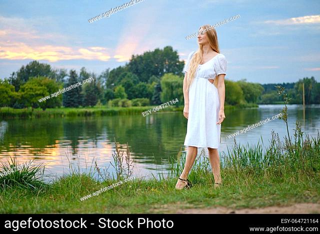 Young woman in white sundress walking along the river bank during sunset