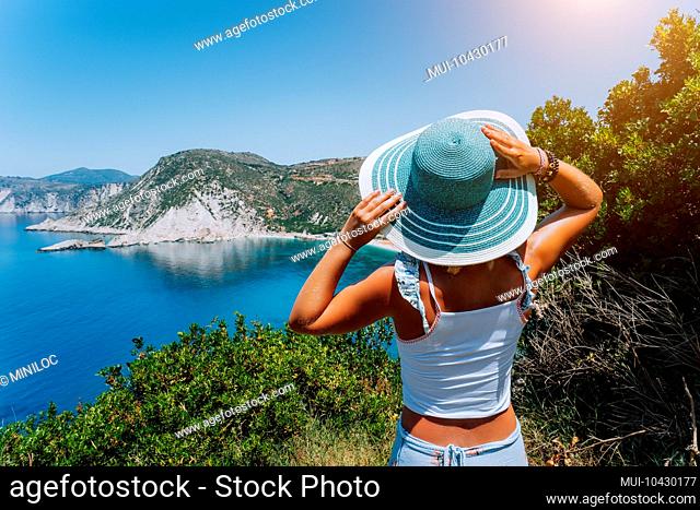 Petani beach Kefalonia. Young woman holding blue sun hat enjoying beautiful panorama of blue bay lagoon surrounded by steep cliff coastline. Greece
