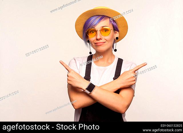 Portrait of glamour positive hipster girl with violet hair in sunglasses and hat standing with crossed hands and pointing to copy space on both sides