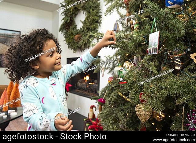 Small girl enjoying decorating and hanging ornaments on Christmas tree in living room at home