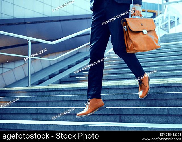 Businessman making steps down stairs. Cropped shot of man wearing elegant suit and shoes walking down stairs and holding brown leather bag in his hand