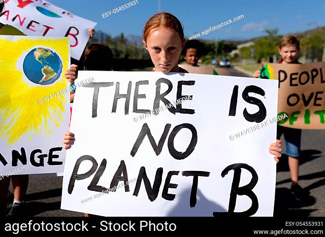 Portrait of a Caucasian girl carrying a sign with a slogan on it and walking down a road in the sun with a diverse group of elementary school pupils also...