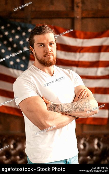 A Successful American Entrepreneur Proudly Poses In His Office Amid American's Flag On Blurred Background. A Bearded European Man In A White T-Shirt Looks...