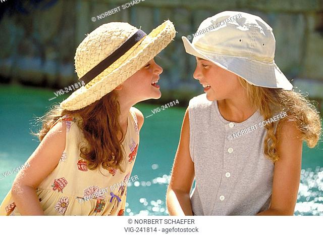 outdoor, half-figure, profile of two laughing 8-10-year-old girls with long hair wearing summerdresses and hats at the border of a swimming pool  - GERMANY