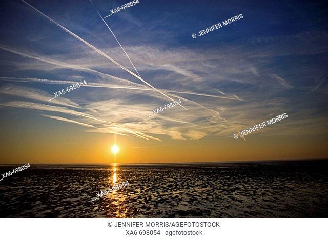 A setting sun creates an orange layer in the deep blue sky and blazes a gold trail across the wet sand of the beach. White vapour trails criss-cross the sky