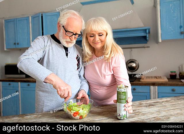Charming elderly couple preparing lunch together. Aged grandpa mixing vegetables in salad bowl with his wife next to him reaching for oil