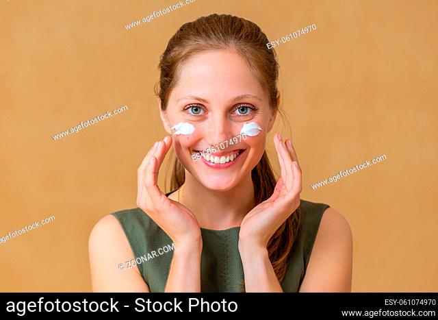 Happy woman applying white cream on her face and looking into camera. Cheerful Caucasian natural looking lady putting on facial cosmetic with hands in yellow...