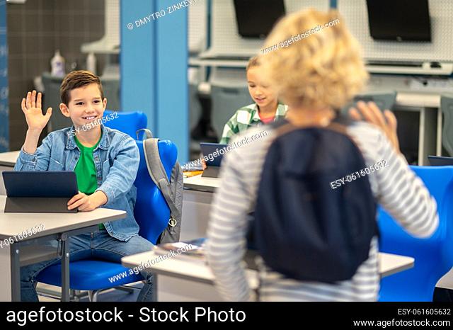Greetings. Light haired boy in backpack standing with back to camera greeting classmate raising hand sitting at desk with tablet