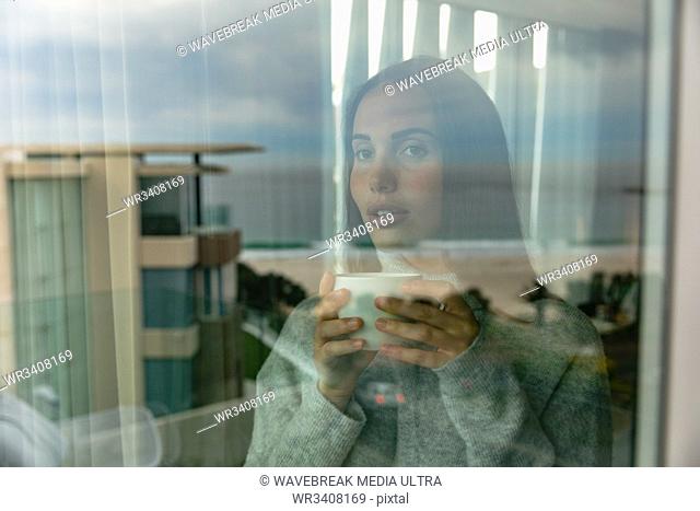 Front view close up of a young Caucasian brunette woman wearing a grey sweater, standing and looking out of a window, holding a cup of coffee