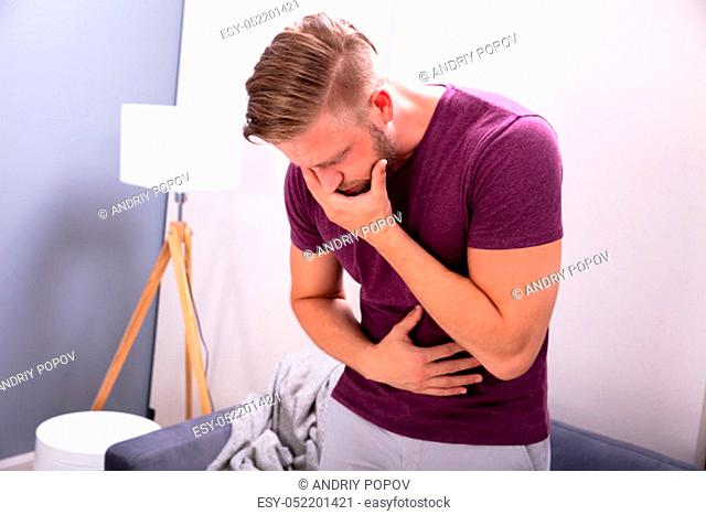 Young Man Suffering From Nausea Standing In Front Of Sofa At Home