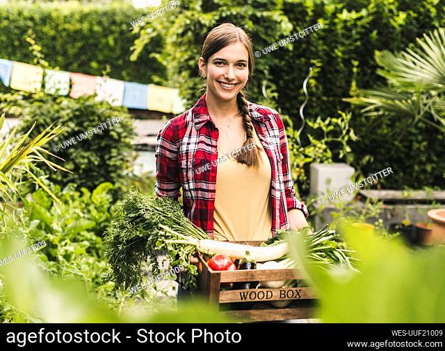 Smiling woman carrying vegetables in crate while standing against plants at garden