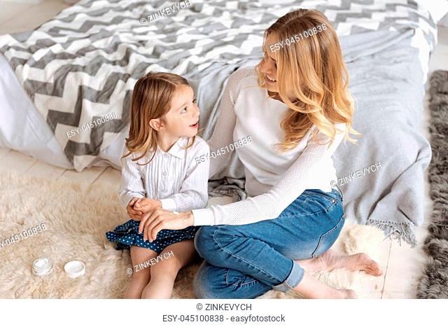 Full of love. Cute little daughter and her beautiful mother sitting on the carpet and looking at each other with smiles while the mother having some cream...
