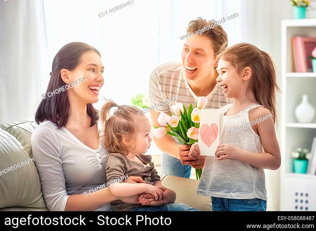 Happy mother's day! Two children daughters with dad congratulate mom and give her a postcard and flowers tulips. Mum and girls smiling and hugging