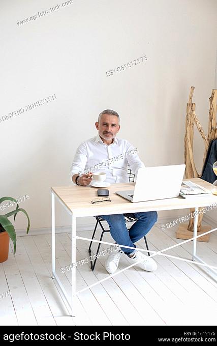 Confidence. Gray-haired business man with coffee sitting at table with laptop looking confidently at camera in daylight room