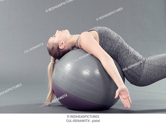 Young woman practising yoga with gym ball, grey background