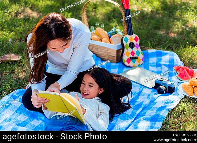 Happy Asian young family mother and child little girl having fun and enjoying outdoor laying on picnic blanket reading book at summer garden spring park