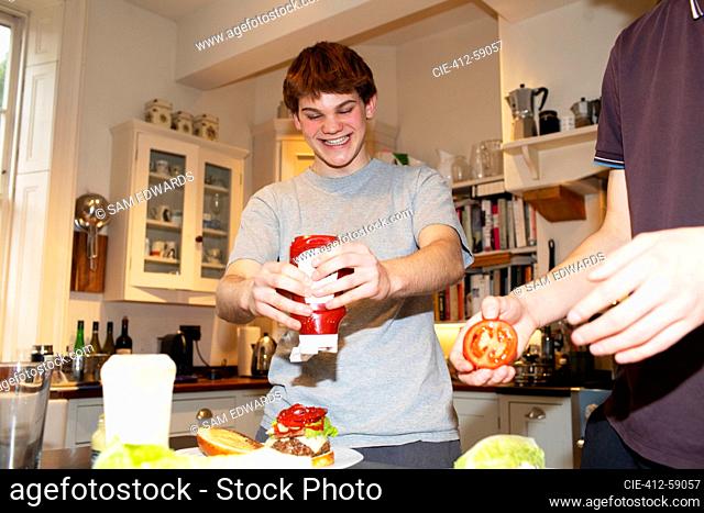 Happy teenage boy squeezing ketchup onto hamburger in kitchen