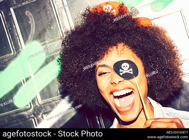 Close-up of cheerful woman holding fake pirate eye patch against wall