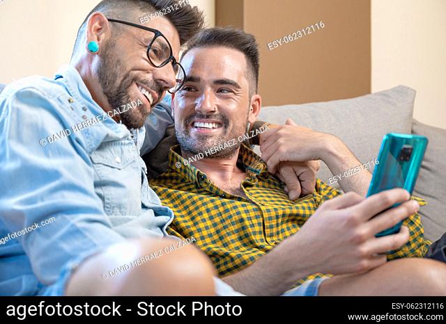 Cheerful gay couple sitting on the couch hugging and looking at the mobile phone, watching a movie, scrolling through the social network