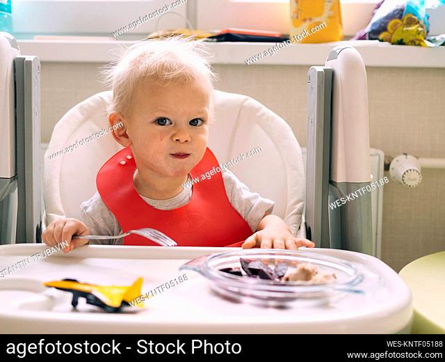 Baby boy sitting on high chair while eating breakfast in kitchen