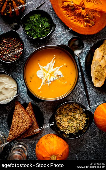 Delicious pumpkin soup with cream, seeds, bread and fresh herbs in elegant ceramic black bowl on dark stone background. Top view flat lay