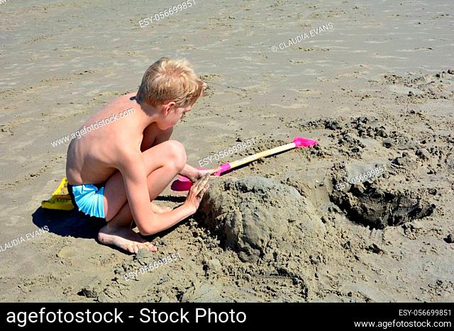 A blond boy with swimming trunks builds a sand castle in the sand on the beach