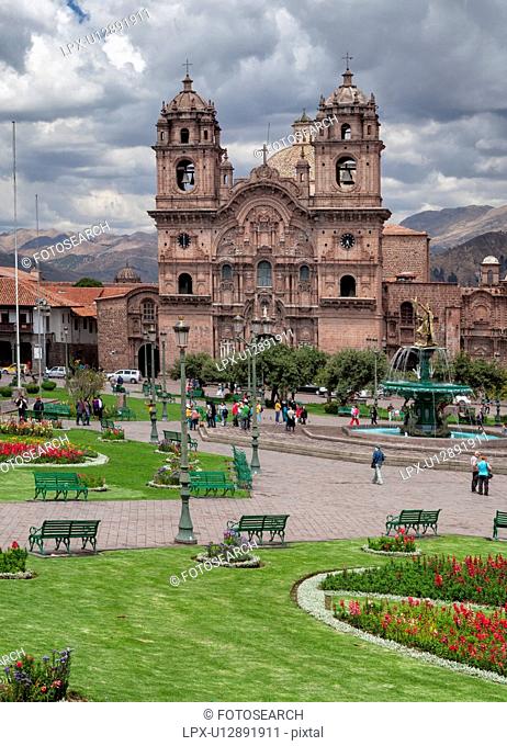 Cuzco: Plaza de Armas, view of Iglesia de la Compania, central fountain with statue, people walking after afternoon rain shower, blue sky with dramatic clouds