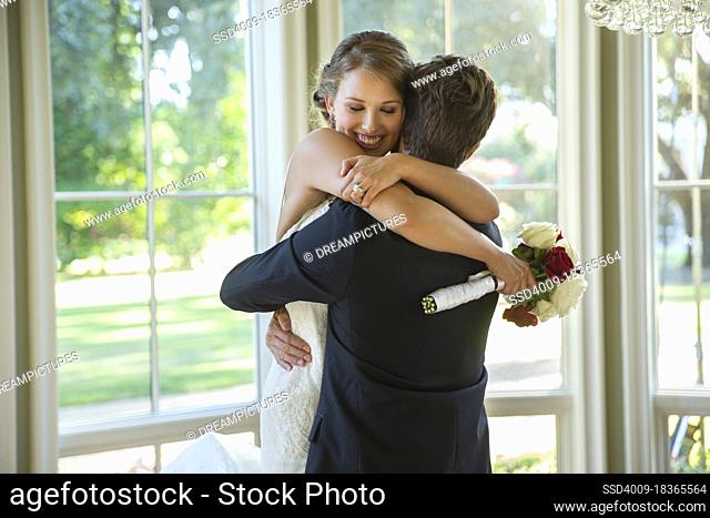 Bride and groom in an embrace, groom lifting up bride standing by large bank of windows