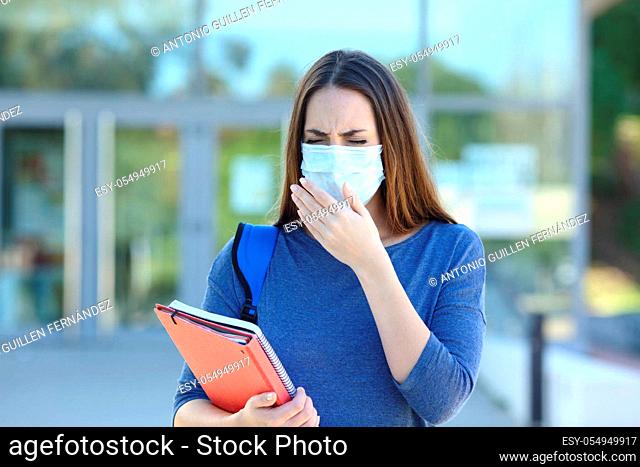 Front view portrait of an ill student wearing a protective mask coughing in the stret