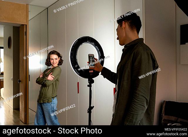 Young man photographing vlogger standing with arms crossed by ring light at home