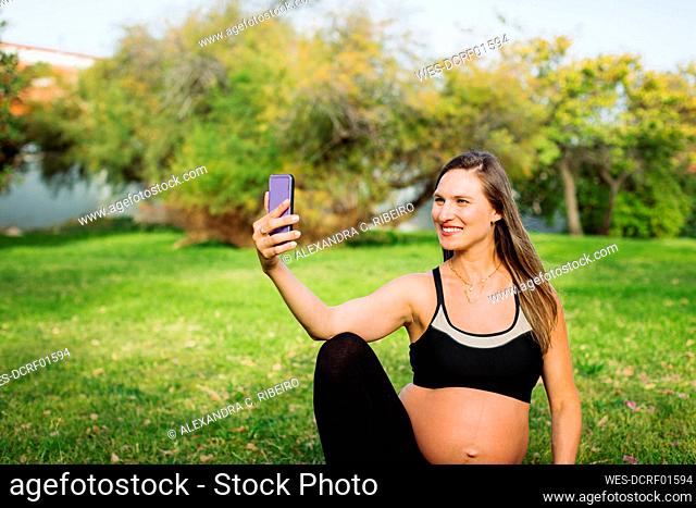 Smiling pregnant woman taking selfie through smart phone sitting on lawn